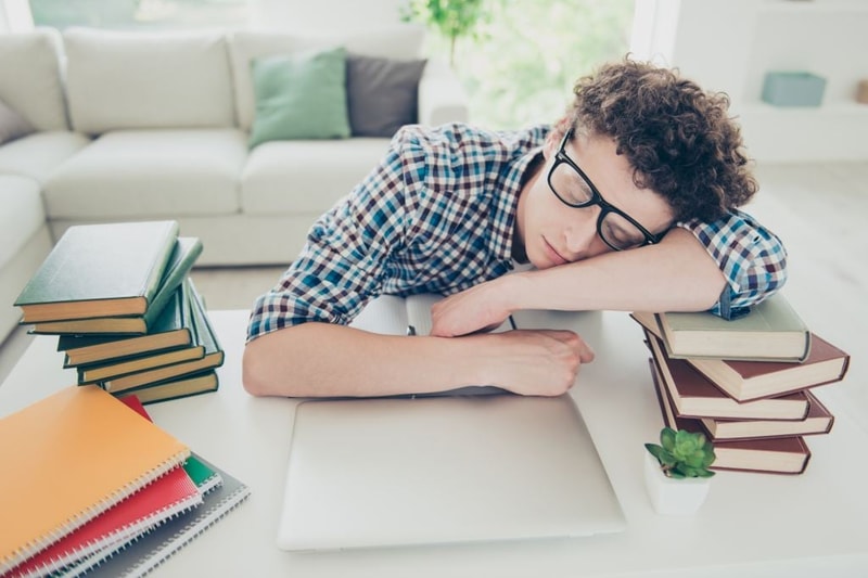 Handsome curly-haired tired young guy student at home, nerd, wearing casual, glasses, sleeping, face and hands on books