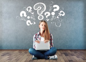 Attractive young woman sitting on wooden floor with laptop and drawn question marks above on concrete wall. Confusion concept