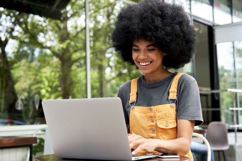Happy hipster African American female student, freelancer with Afro hair using laptop computer sitting at table in outdoor cafe elearning, remote studying, typing on pc tech, working on internet.