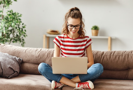 Cheerful young female freelancer in glasses and casual clothes focusing on screen and interacting with laptop while sitting alone on soft couch in light modern living room