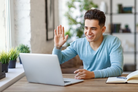 Happy ethnic guy smiling and waving hand while sitting at table and having online lesson at home
