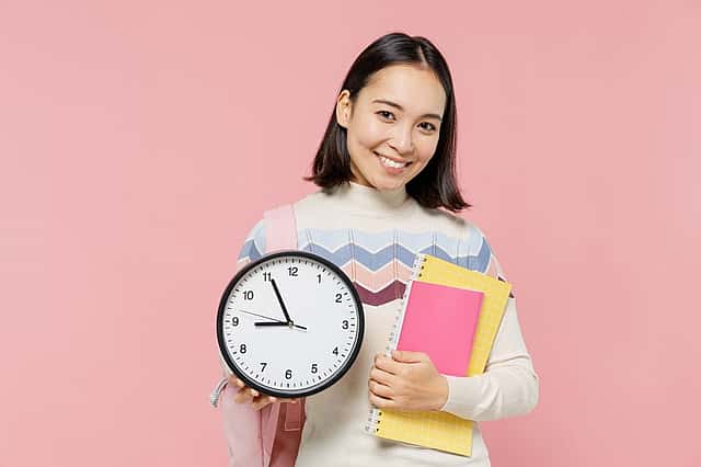 Smiling happy satisfied teen student girl of Asian ethnicity wear sweater backpack hold books clock isolated on pastel plain light pink background Education in high school university college concept