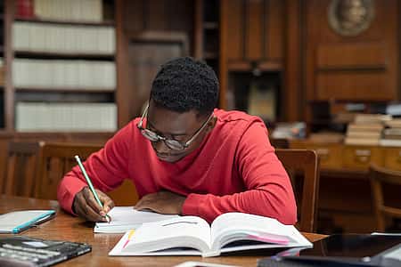 University student wearing spectacles studying in library. Young african man taking notes from book while sitting in the library. Focused casual guy writing notes during homework.