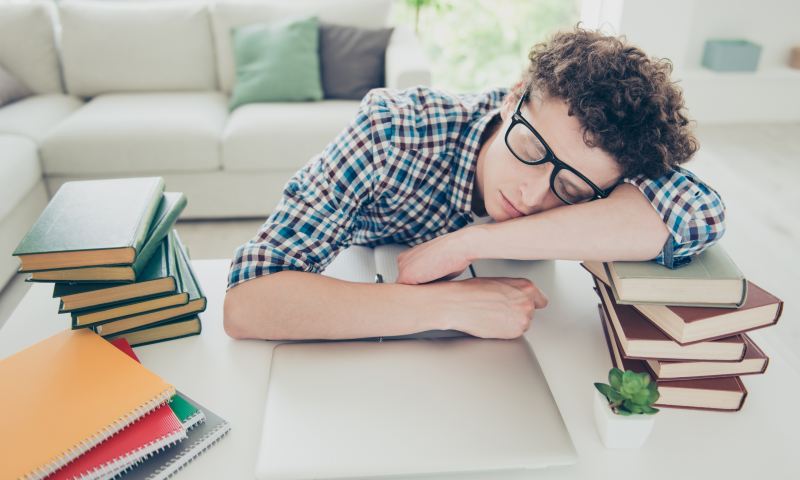 Handsome curly-haired tired young guy student at home, nerd, wearing casual, glasses, sleeping, face and hands on books