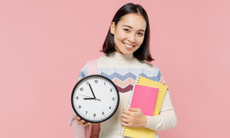 Smiling happy satisfied teen student girl of Asian ethnicity wear sweater backpack hold books clock isolated on pastel plain light pink background Education in high school university college concept