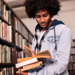 African male college student standing in library with lots of books. University student looking for study references.