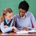 Young African American female teacher assisting schoolgirl at desk in classroom