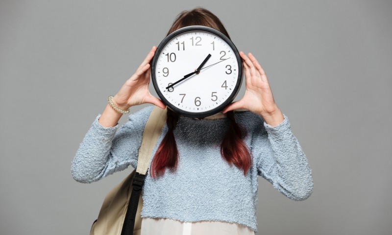 Image of girl standing isolated grey background covering face with clock in hands.