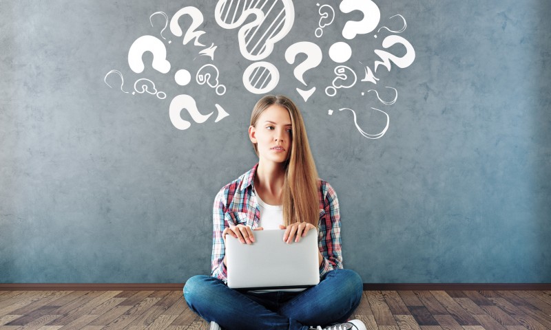 Attractive young woman sitting on wooden floor with laptop and drawn question marks above on concrete wall. Confusion concept