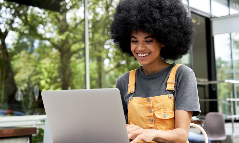 Happy hipster African American female student, freelancer with Afro hair using laptop computer sitting at table in outdoor cafe elearning, remote studying, typing on pc tech, working on internet.