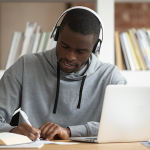 Focused young african american male student sitting at table with computer, wearing bluetooth earphones, busy with study, listening to educational online lecture or enjoying favorite music tracks.
