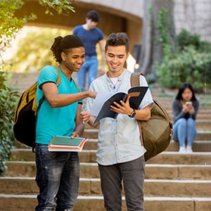 Two young male multiracial students looking at and pointing at a notebook on some stairs with other students behind them.