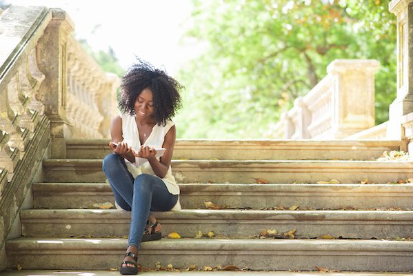 Portrait of happy young African-American female student wearing blouse and jeans sitting on stairs and reading