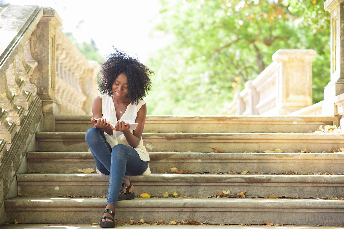 Portrait of happy young African-American female student wearing blouse and jeans sitting on stairs and reading copybook in autumn. Education and leisure concept