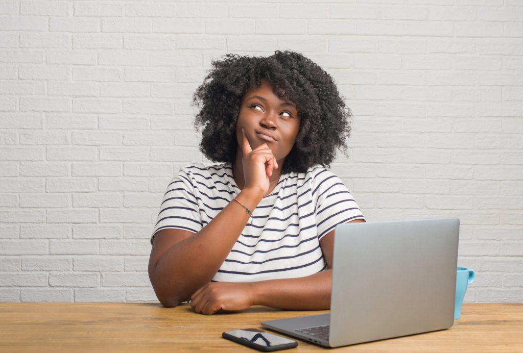 Young african american woman sitting at a table using laptop serious face thinking about question