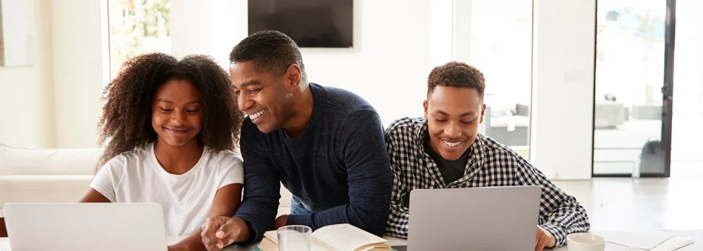 African-American father standing between a boy and a girl with laptops, looking at the girl's laptop