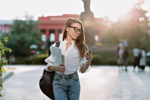 Smiling student checks phone while holding notebook on campus.