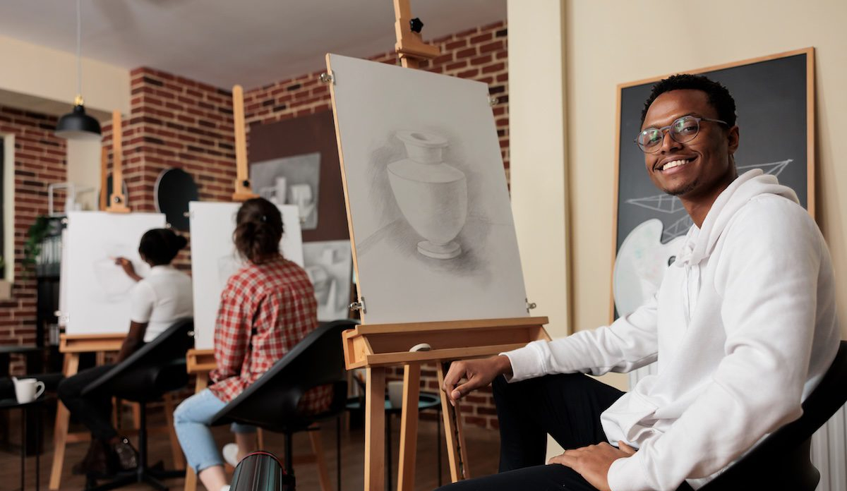 Portrait of happy young African American man student of art school sitting at easel smiling at camera, learning pencil drawing in class. Positive black guy enjoying creative hobby with friends