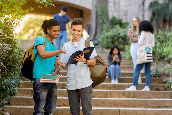 Two young male multiracial students looking at and pointing at a notebook on some stairs with other students behind them.